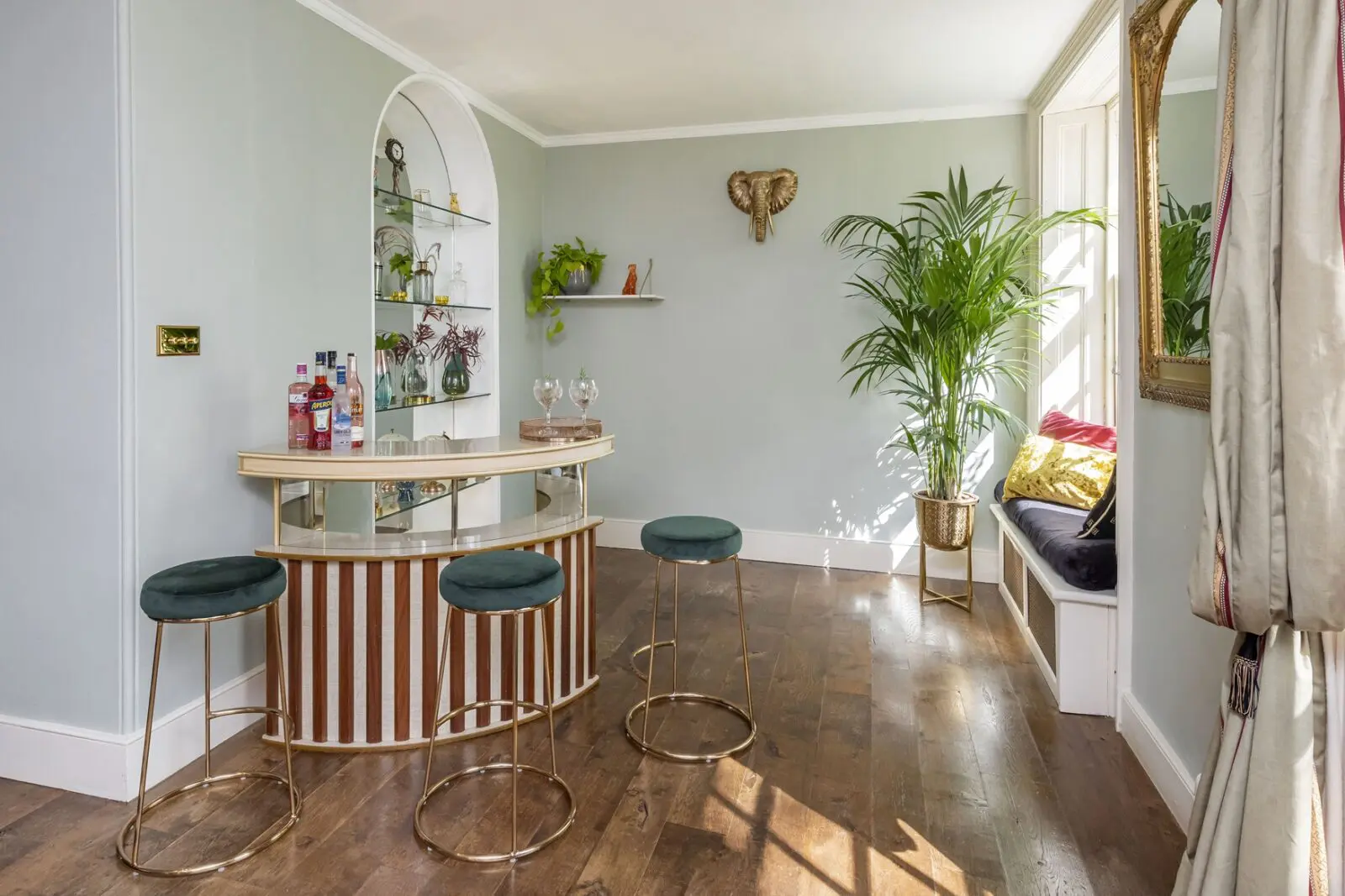A home bar with three green stools, a curved counter, glass shelves with bottles and glasses, potted plants, wooden floor, and a window seat with cushions.