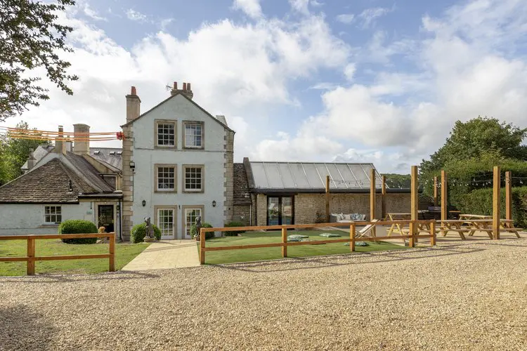 A large light-coloured house with a modern glass extension, outdoor seating, and a gravel area under a partly cloudy sky.