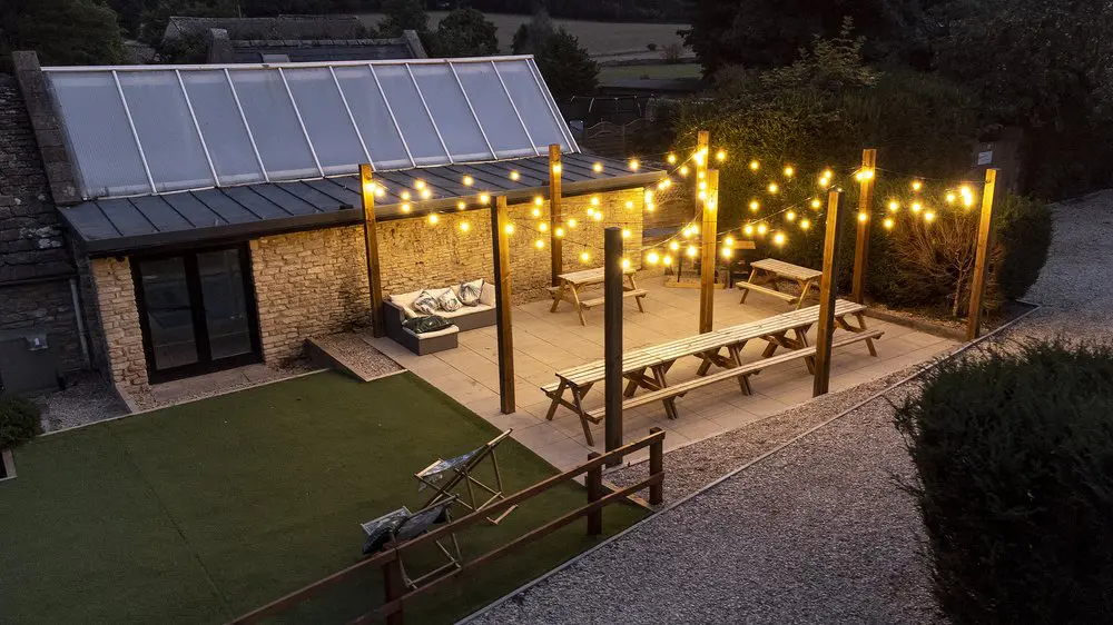 Outdoor patio with fairy lights, long wooden tables, benches, a small artificial grass area, and a stone building in the background, viewed at dusk.