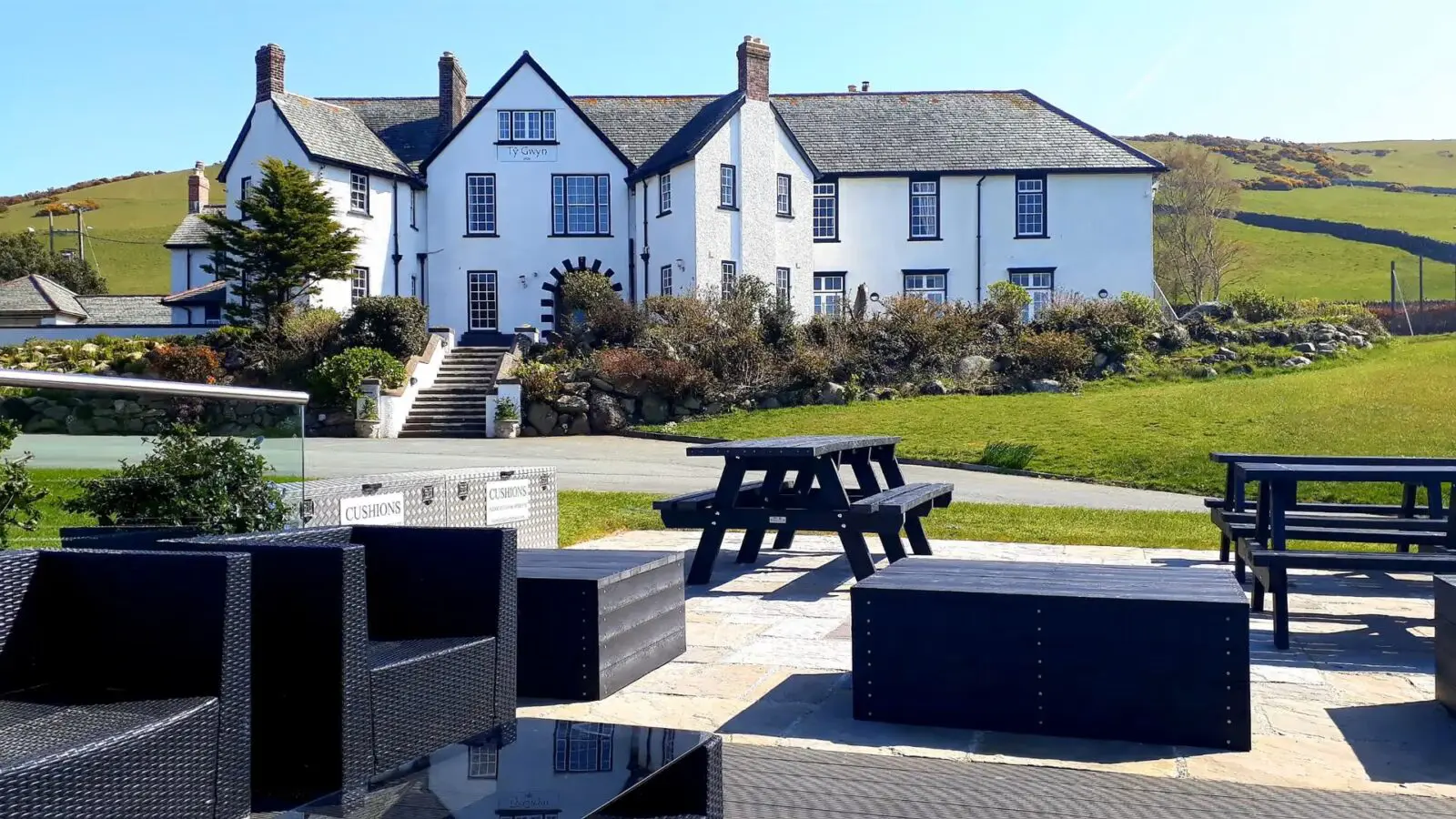 Large white country house with multiple chimneys, set on a hill. Outdoor patio in the foreground features black picnic tables and wicker chairs. Green lawns and hills surround the building.