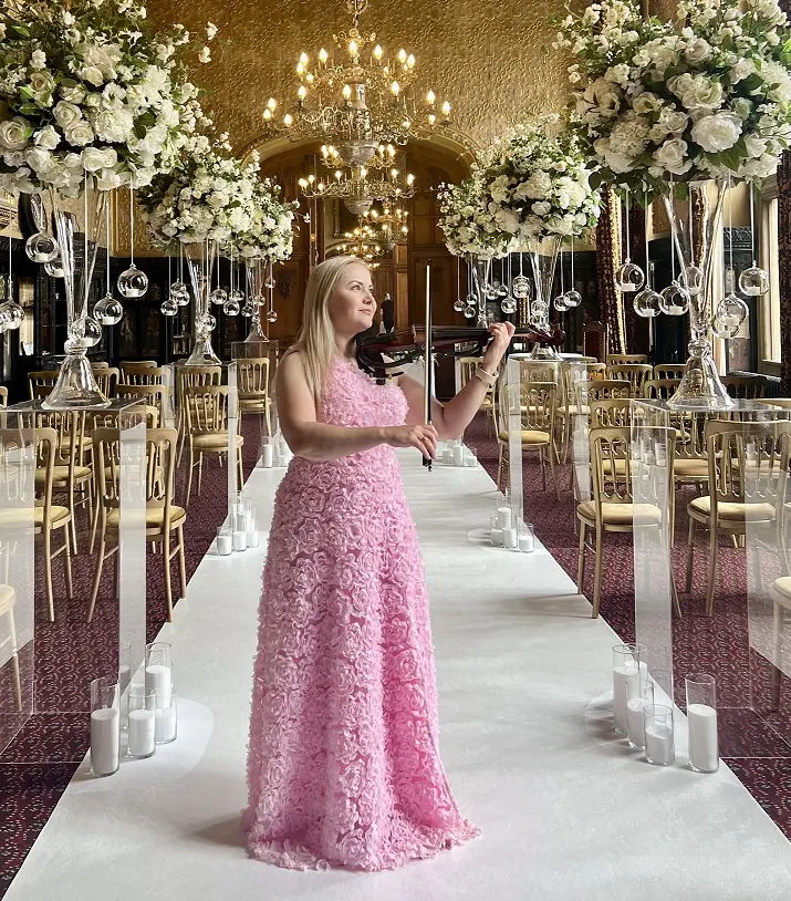 A woman in a pink lace dress plays the violin in an ornate hall decorated with white flowers, chandeliers, and rows of chairs.