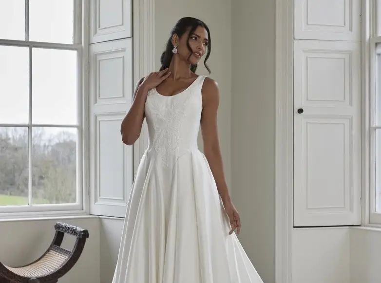 A woman in a sleeveless white wedding dress stands indoors near tall windows, holding part of her dress and looking to the side.