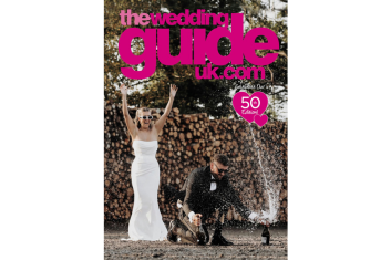 A bride in a white dress raises her arms while a groom in a tuxedo opens a bottle of champagne, splashing liquid, in front of stacked logs. “The Wedding Guide UK” text is above them.