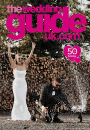 A bride and groom celebrate outdoors; the groom pops a bottle of champagne as the bride raises her arms. Text reads “the wedding guide uk.com” and “50th edition”.