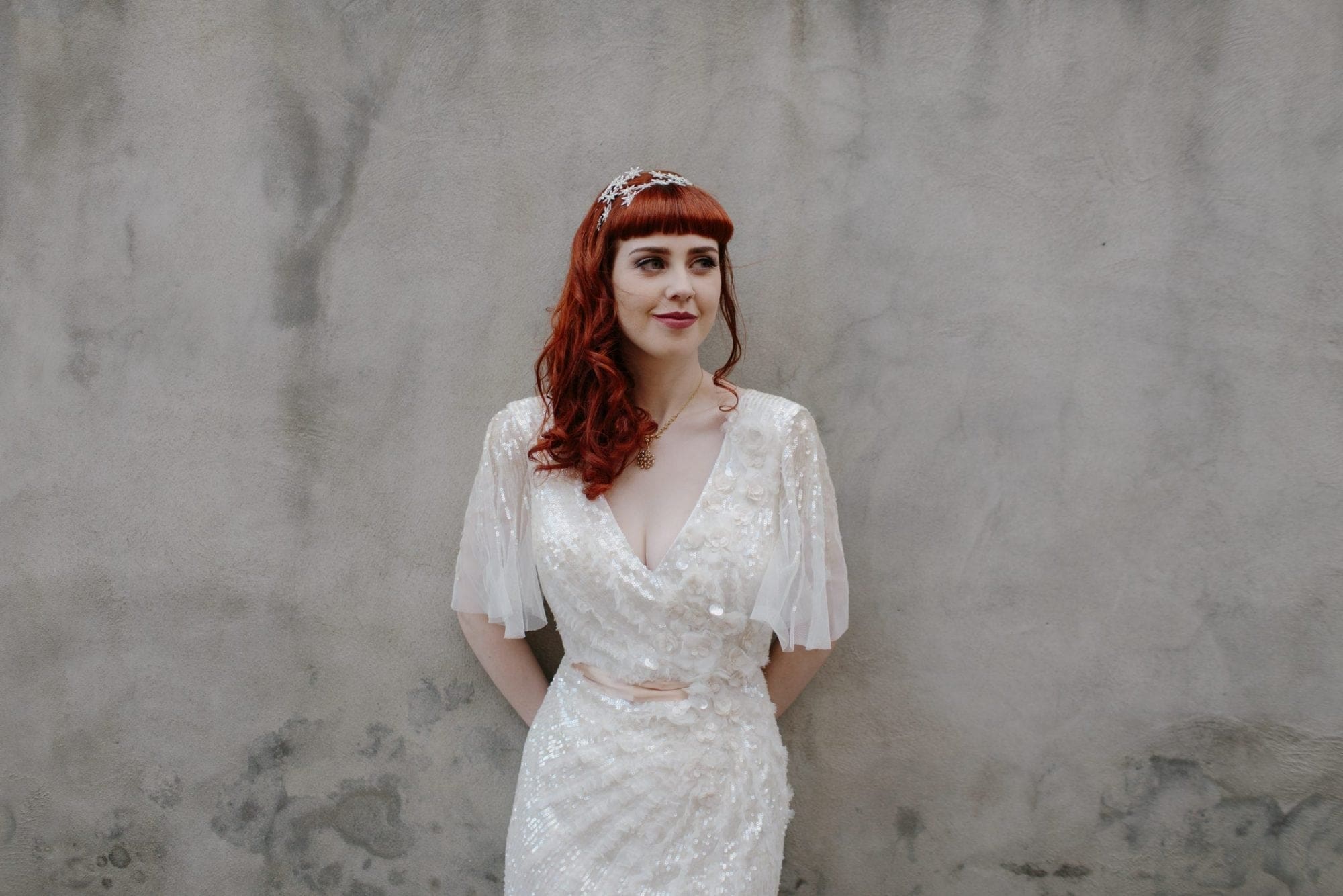 A red haired woman in a white wedding dress leaning against a wall on her special day.
