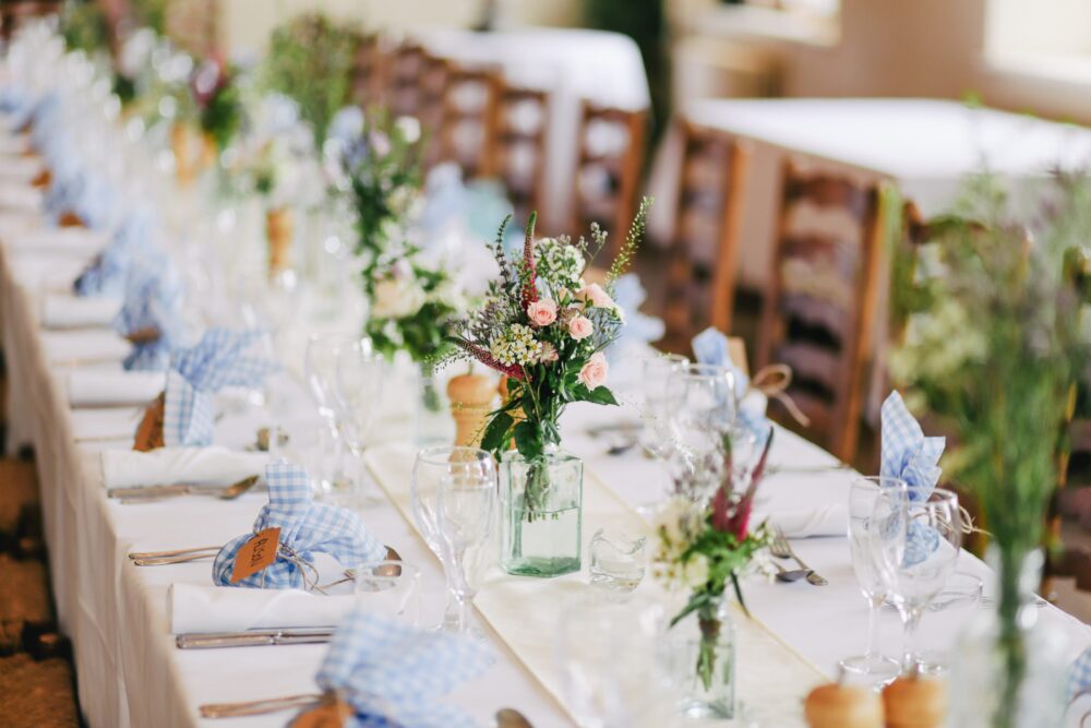 A long dining table is set with white linens, glassware, and floral centerpieces. Napkins are folded and tied with blue ribbon. Wooden chairs line either side of the table.