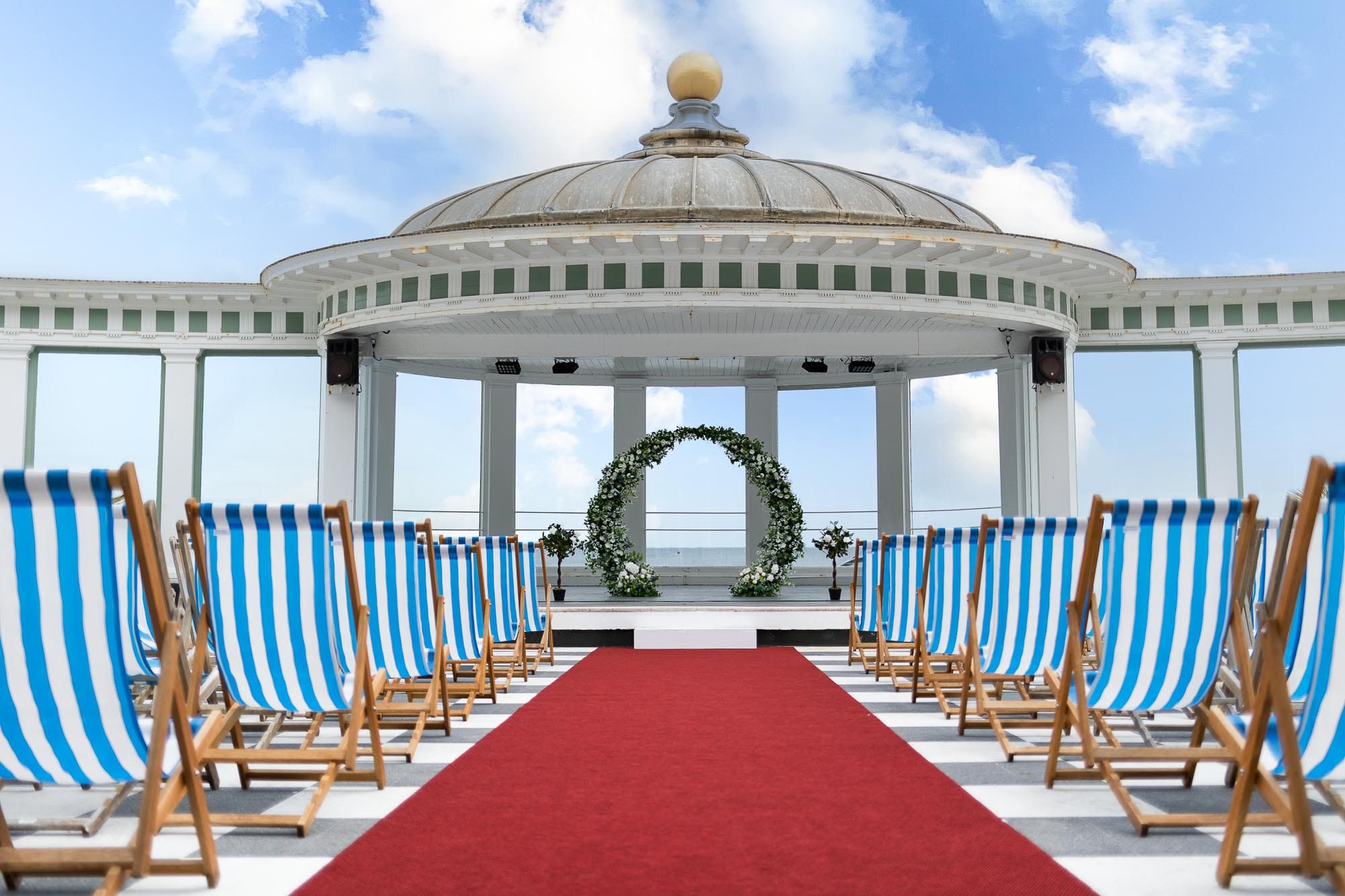 An enchanting outdoor wedding venue featuring striped deck chairs facing an altar under a gazebo adorned with a floral arch. A red carpet elegantly leads to the altar, set against a breathtaking blue sky. Perfect for those seeking premium wedding services in the UK.