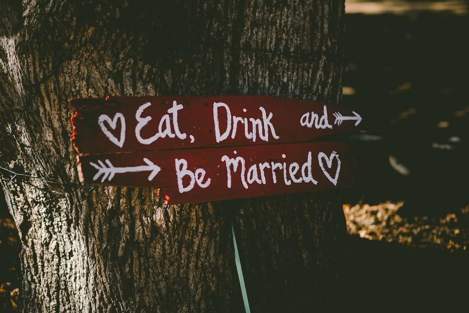 Handwritten sign attached to a tree saying "eat, drink and be married" with directional arrows and heart symbols.