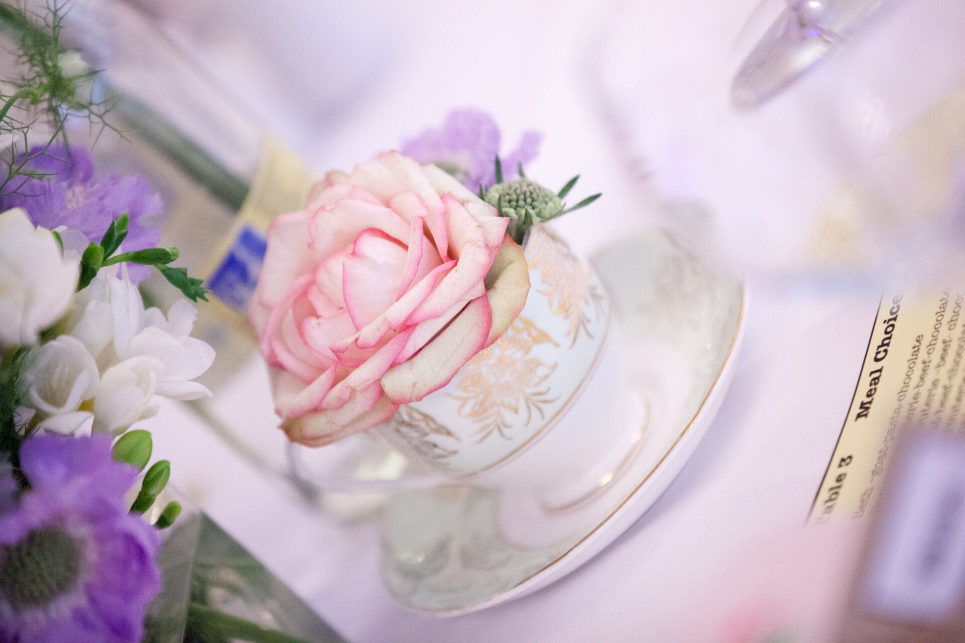A close-up of a pink rose arranged in a white, gold-trimmed teacup surrounded by other flowers and a blurred background.